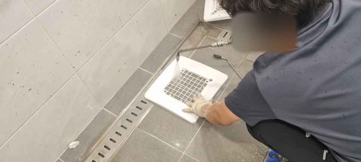 회색 타일 바닥에 쪼그려 앉아 장갑 낀 손으로 흰 격자 틀을 누르고 세척기로 물을 쏘는 작업자